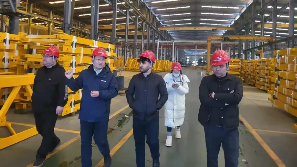 Five-member tour group in red safety helmets walks through the steel-beam storage bay at Shuyue's Liangshan plant under an EUROCRAINE overhead crane